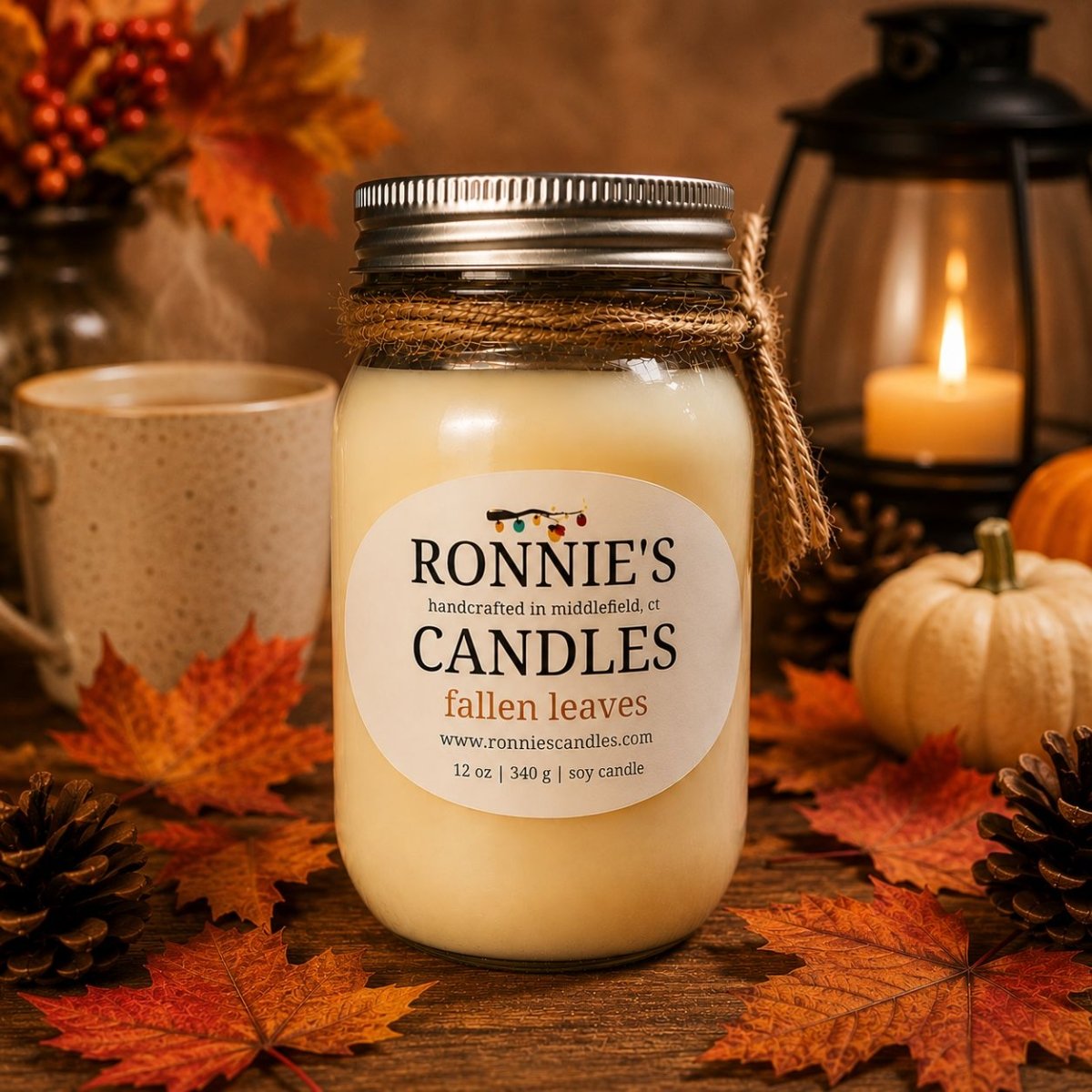 A jar candle labeled 'RONNIE'S CANDLES fallen leaves' sits on a wooden surface surrounded by autumn leaves, pinecones, a mug, pumpkins, and a lantern with a lit candle in the background.