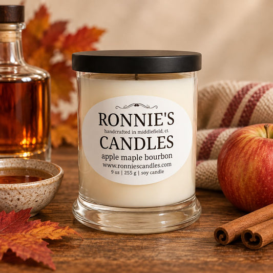A jar candle labeled 'RONNIE'S CANDLES apple maple bourbon' sits on a wooden table with autumn-themed props including an apple, cinnamon sticks, a small bowl, and a bottle of bourbon, against a warm neutral background.