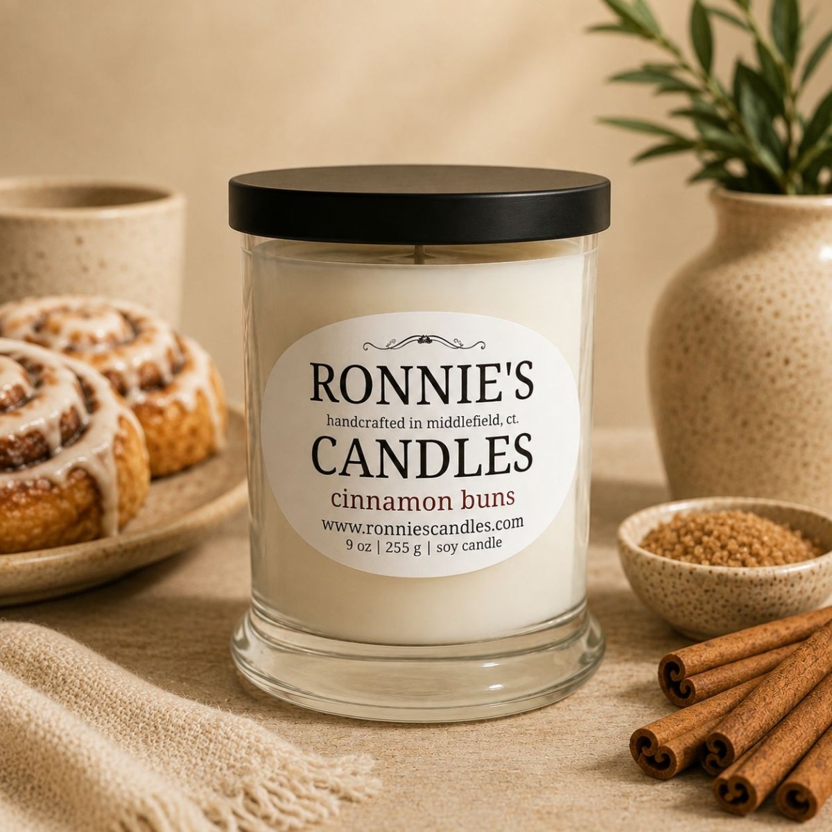 A 9 oz soy candle in a clear glass jar with a black lid and a white label reading 'RONNIE'S CANDLES' and 'cinnamon buns'. The candle is styled on a beige fabric surface with cinnamon sticks, a small bowl of brown sugar, a plate of cinnamon rolls, and ceramic vessels in the background.