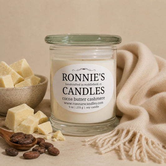 A glass jar candle labeled 'RONNIE'S Candles cocoa butter cashmere' sits on a beige surface with cream-colored wax blocks, cocoa beans, and a cream knit throw in a warm styled setting.