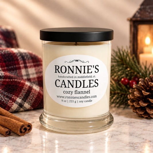 A 9 oz soy candle in a clear glass jar with a black lid and a white label reading 'RONNIE'S CANDLES cozy flannel'. The candle is styled on a marble surface with a plaid blanket, cinnamon sticks, pine branches, and a lantern in the background.