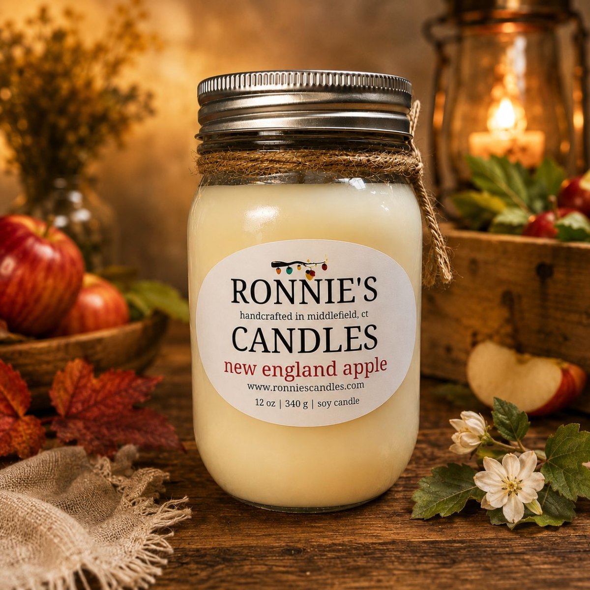 A mason jar candle labeled 'RONNIE'S CANDLES new england apple' sits on a rustic wooden table surrounded by apples, leaves, flowers, and a warm lantern in the background.