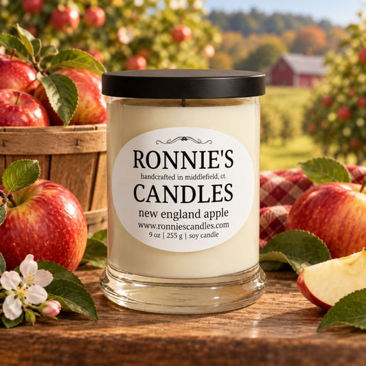 A 9 oz soy candle in a clear glass jar with a black lid and a white label reading 'RONNIE'S CANDLES', set on a wooden table with red apples, green leaves, and a blurred outdoor apple orchard background.