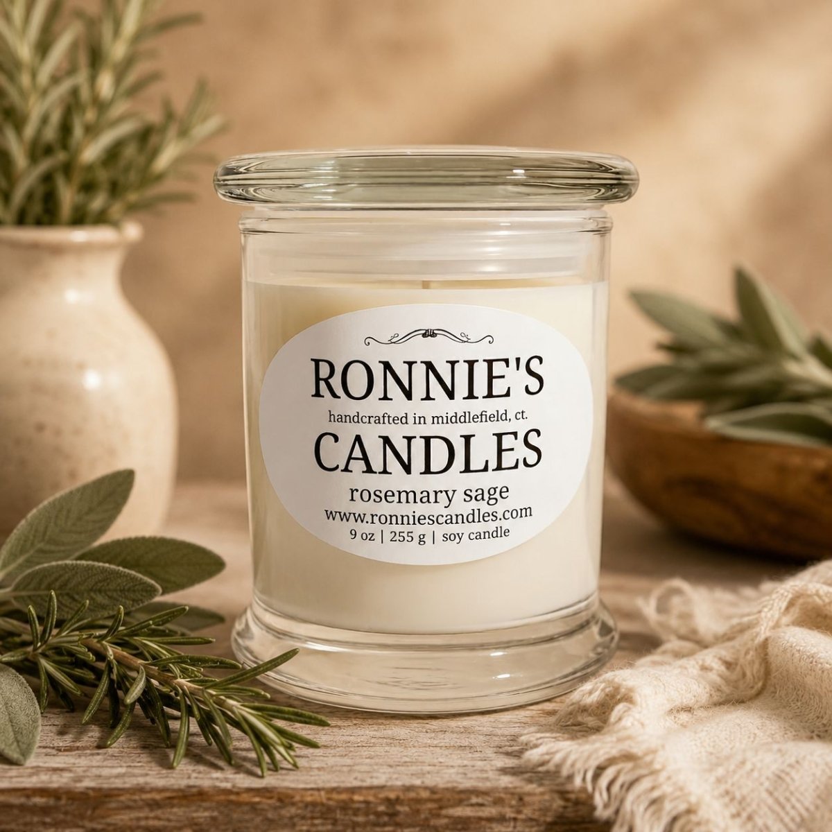 A clear glass jar candle with white wax sits on a rustic wooden surface. The front label reads 'RONNIE'S CANDLES' and 'rosemary sage.