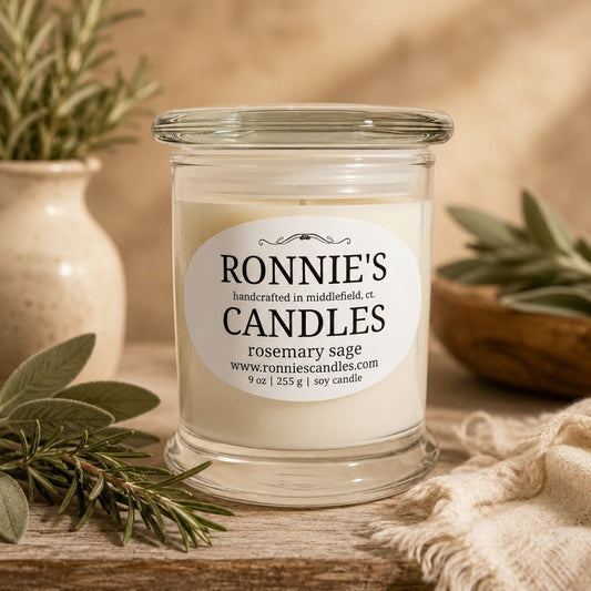 A clear glass jar candle with white wax sits on a rustic wooden surface. The front label reads 'RONNIE'S CANDLES' and 'rosemary sage.